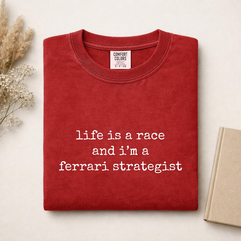 Op de afbeelding: Een rood t-shirt met ronde hals en de witte tekst "life is a race and i'm a ferrari strategist" op de voorkant. Het shirt heeft een Comfort Colors merklabel aan de binnenkant van de kraag en is gemaakt van een effen rode stof.