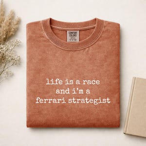 Op de afbeelding: Een terracotta-kleurig t-shirt met ronde hals en een vintage gewassen textuur. Het shirt is voorzien van de witte opdruk "life is a race and i'm a ferrari strategist" in het midden op de borst en een Comfort Colors label in de nek.