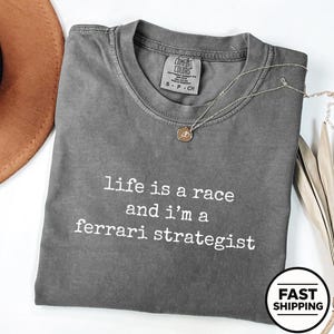 Op de afbeelding: Een antracietgrijs t-shirt met korte mouwen, een ronde hals en een witte opdruk met de tekst "life is a race and i'm a ferrari strategist". Het shirt is voorzien van een Comfort Colors merklabel in de nek met de maat S.