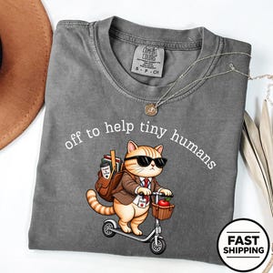 Puede incluir: Una camiseta gris carbón con un gráfico de un gato atigrado naranja con gafas de sol, blazer marrón y corbata roja, montado en un patinete. El gato lleva una mochila con libros y una cesta con una manzana. El texto dice: off to help tiny humans.