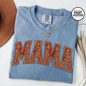 Può includere: Una t-shirt blu a maniche corte con scollo rotondo che presenta la parola MAMA in grandi lettere arcuate rosso scuro con un motivo floreale dorato. La maglietta ha un'etichetta del marchio Comfort Colors sul collo e una piccola collana dorata.