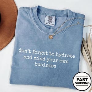 Op de afbeelding: Een blauw T-shirt met korte mouwen, een ronde hals en een Comfort Colors merklabel. Het shirt is voorzien van de witte tekst: don't forget to hydrate and mind your own business. Een goudkleurige ketting met een kleine ronde hanger ligt erbovenop.