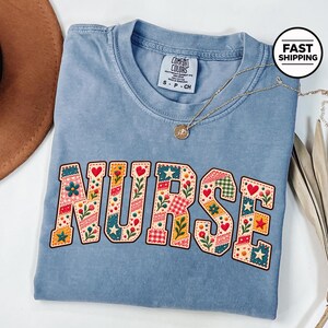 Op de afbeelding: Een blauw T-shirt met korte mouwen en een ronde hals, voorzien van het woord NURSE in grote blokletters. De letters zijn gevuld met een kleurrijk patchworkpatroon van bloemen, hartjes, sterren en rood-witte ruitjes in rood, groen en geel.