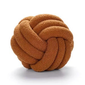 Op de afbeelding: Een bolvormig knoopkussen gemaakt van een gestructureerde, pluizige bruine stof. Het ontwerp bestaat uit een doorlopende, verweven lus die een ronde, balachtige vorm cre&euml;ert met een zacht aanvoelend oppervlak en een uniforme, warme bruine kleur.
