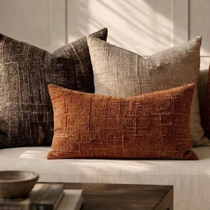 May include: A collection of textured throw pillows in various earth tones. The pillows are arranged on a white sofa, with a focus on a rectangular, rust-colored pillow with a grid pattern. Other pillows are in shades of brown and beige.