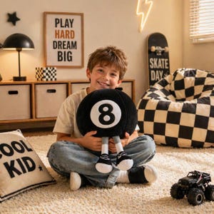 Op de afbeelding: Een zwart en wit 8-ball kussen met bevestigde benen en sneakers. Het kussen wordt door een persoon vastgehouden. Een kussen met de woorden "COOL KID" staat op de voorgrond. Een ingelijst bord luidt "PLAY HARD DREAM BIG".