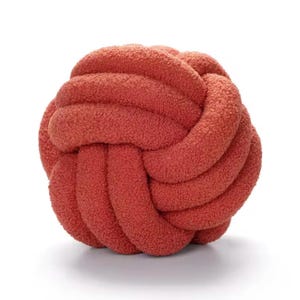 Op de afbeelding: Een bolvormig knoopkussen gemaakt van een getextureerde, pluizige stof in een terracotta kleur. Het ontwerp bestaat uit een doorlopende, verweven buis die een ronde, balachtige vorm vormt met een zacht oppervlak en een consistente diepe oranjerode tint.