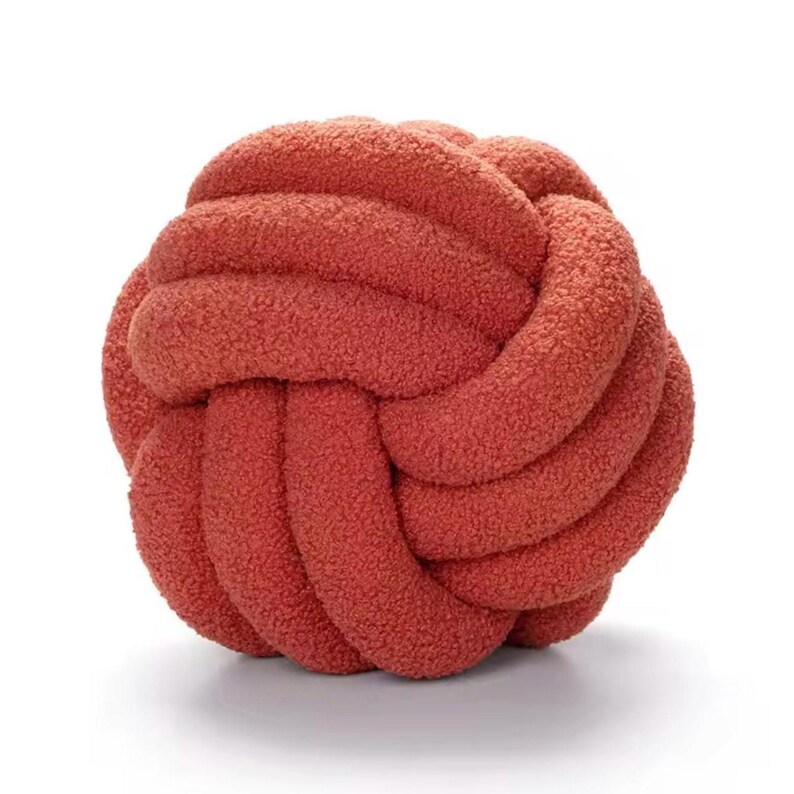 Op de afbeelding: Een bolvormig knoopkussen gemaakt van een getextureerde, pluizige stof in een terracotta kleur. Het ontwerp bestaat uit een doorlopende, verweven buis die een ronde, balachtige vorm vormt met een zacht oppervlak en een consistente diepe oranjerode tint.