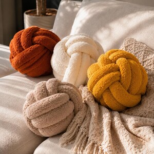Op de afbeelding: Vier ronde knoopkussens gemaakt van gestructureerde, pluche stof in de kleuren gebrand oranje, cr&egrave;me, off-white en mosterdgeel. Elk kussen heeft een doorlopend, verweven ontwerp dat een bolvorm cre&euml;ert met een zacht, pluizig oppervlak.