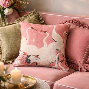 May include: A square decorative pillow with a pink background featuring a white swan design. The pillow is on a pink sofa with other pillows in shades of green and pink. A gold tray with a lit candle is in the foreground.
