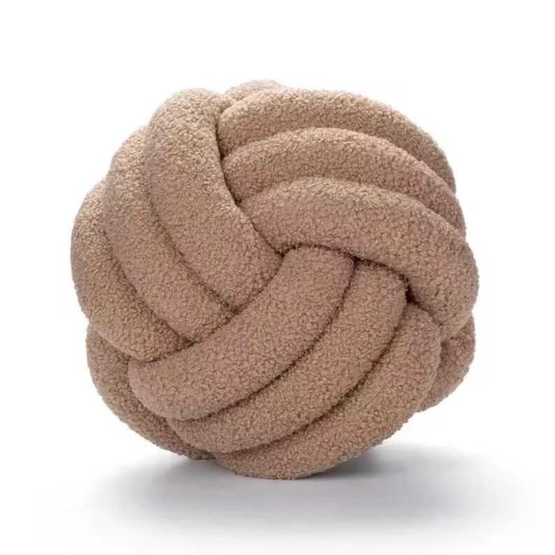 Op de afbeelding: Een bolvormig knoopkussen gemaakt van een zachte, getextureerde, beige stof. Het ontwerp bestaat uit een doorlopende, verweven lus die een complexe, ronde vorm cre&euml;ert met een pluizige, fleece-achtige oppervlaktestructuur over het object.