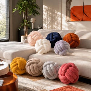 Op de afbeelding: Een verzameling van negen ronde knoopkussens gemaakt van zachte, getextureerde stof in diverse kleuren, waaronder lichtroze, marineblauw, roestoranje, wit, lavendel, mosterdgeel, beige, lichtgrijs en koraalroze, gerangschikt op een bank.