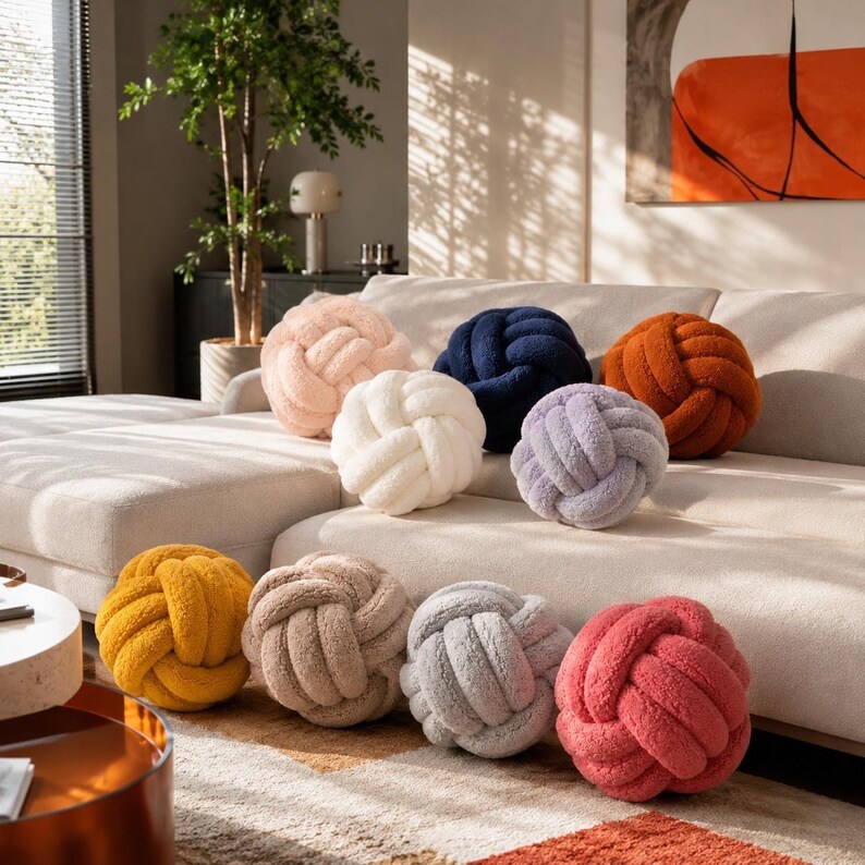 Op de afbeelding: Een verzameling van negen ronde knoopkussens gemaakt van zachte, getextureerde stof in diverse kleuren, waaronder lichtroze, marineblauw, roestoranje, wit, lavendel, mosterdgeel, beige, lichtgrijs en koraalroze, gerangschikt op een bank.