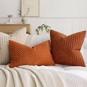 Op de afbeelding: Drie decoratieve kussens in crème- en oranjetinten liggen op een witte bank. De oranje kussens hebben een gestructureerd stippenpatroon. De bank is bedekt met een witte, gebreide deken.