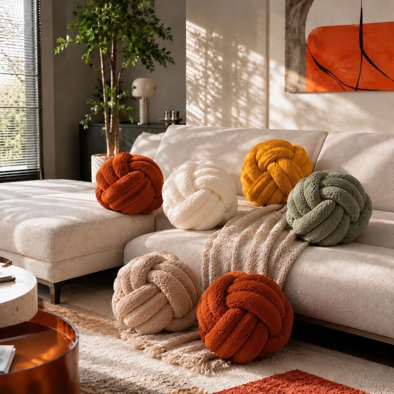 Op de afbeelding: Een verzameling van zes bolvormige knoopkussens in diverse kleuren, waaronder gebrand oranje, cr&egrave;me, mosterdgeel en saliegroen. Elk kussen heeft een textuurrijk, pluche stoffen oppervlak dat in een complex knoopontwerp is geweven.