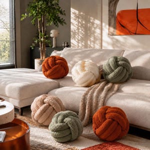Op de afbeelding: Een collectie van zes bolvormige knoopkussens gemaakt van textuurrijke stof in de kleuren gebrand oranje, cr&egrave;me, beige en saliegroen. Deze ronde, geknoopte kussens zijn gerangschikt op een lichte bank en een vloerkleed en hebben een zacht oppervlak.