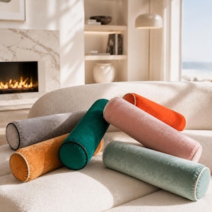 May include: A selection of cylindrical cushions in shades of orange, pink, green, and grey, arranged on a cream sofa. The cushions have a soft, velvety texture and are set against a backdrop of a fireplace and shelves. The cushions are a decorative home accessory.