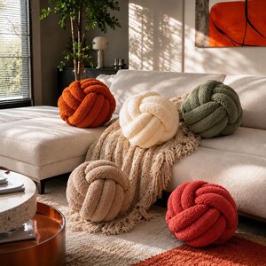 Op de afbeelding: Vijf bolvormige knoopkussens van gestructureerde, pluche stof in de kleuren gebrand oranje, cr&egrave;me, olijfgroen, beige en rood. Deze ronde, geknoopte kussens zijn gerangschikt op een lichtgekleurde bank, met &eacute;&eacute;n cr&egrave;mekleurig kussen op een plaid met franjes.