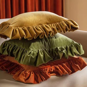 May include: Three square velvet cushions in gold, olive green, and burnt orange. Each cushion has a ruffled edge. The cushions are stacked, creating a layered effect. The image shows a close-up of the cushions.