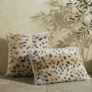 May include: Two faux fur throw pillows with a spotted pattern in shades of beige, cream, and brown. The pillows are square and rectangular, with a soft, plush texture. The pillows are designed for home decor.