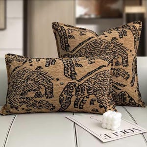 May include: Two decorative cushions with a tan background and a black pattern. The cushions are rectangular and square, with a detailed design. A white candle and a magazine are in the foreground.