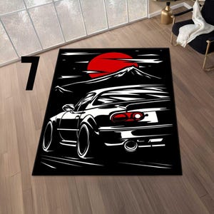 May include: Black and white area rug featuring a stylized illustration of a sports car with a red sun and mountain range in the background. The rug has a rectangular shape and is placed on a wooden floor.