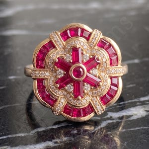 May include: A gold ring featuring a central round ruby surrounded by six baguette-cut rubies in a star pattern, accented by small round diamonds and an outer border of baguette-cut rubies arranged in a scalloped, floral-inspired circular design.