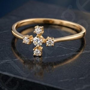 Puede incluir: Un anillo de oro con un adorno central en forma de cruz compuesto por cuatro piedras pequeñas, redondas y transparentes dispuestas en un patrón vertical y horizontal, con una piedra central más grande en la intersección de la cruz.