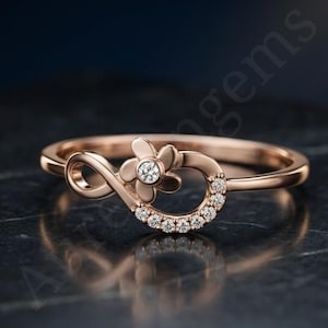 Könnte beinhalten: Ein Ring aus Roségold mit einem Unendlichkeitssymbol, das mit einer kleinen fünfblättrigen Blume und einer Reihe winziger, klarer, runder Steine an einer Seite der Schlaufe verziert ist. Der Ring ist dünn und poliert und zeigt eine feine Verarbeitung.