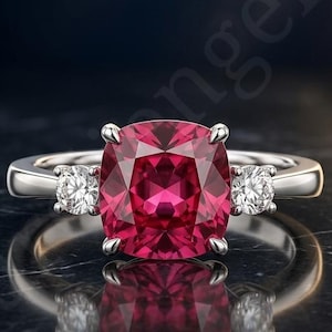 Könnte beinhalten: Ein silberner Ring mit einem großen, tiefrosa Edelstein im Kissenschliff in der Mitte, flankiert von zwei kleineren, runden, klaren weißen Steinen auf beiden Seiten. Das Metallband ist poliert und reflektierend mit glänzendem Finish.