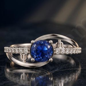 Pode incluir: Um anel de tom prateado com uma pedra central redonda azul-profundo presa por quatro garras. A banda é acentuada com pequenas pedras claras em pavé e incorpora dois símbolos distintos em forma de tridente na estrutura metálica.