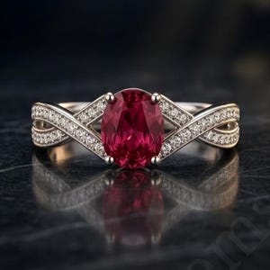Könnte beinhalten: Ein Ring mit einem zentralen, oval geschliffenen, tiefroten Edelstein, der von vier Krappen gehalten wird. Die Ringschiene besteht aus Metall mit einem gekreuzten Design an beiden Seiten, besetzt mit kleinen klaren Steinen. Text Aureliengems.