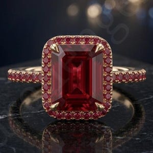 Puede incluir: Un anillo de oro con una gran piedra preciosa de color rojo intenso en talla esmeralda en el centro, rodeada por un halo de piedras rojas más pequeñas. La banda también está incrustada con una fila continua de pequeñas piedras rojas.