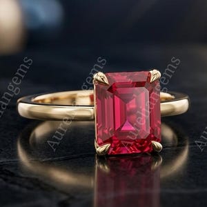 Könnte beinhalten: Ein Goldring mit einem leuchtend roten Edelstein im Smaragdschliff, der von vier Krappen gehalten wird. Die Ringschiene ist aus glattem, poliertem Gelbgold. Der Text Aureliangems ist im Bild sichtbar. Das Design betont die tiefrote Farbe und die Facetten des zentralen Steins, der sich auf der dunklen, strukturierten Oberfläche spiegelt.