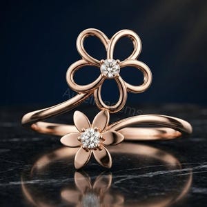 Könnte beinhalten: Ein Bypass-Ring aus Roségold mit zwei Blumenmotiven. Die größere Blume hat offene Blütenblätter mit einem runden, weißen Edelstein in der Mitte, während die kleinere Blume massive Blütenblätter und einen passenden runden, weißen Edelstein besitzt.