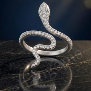 Könnte beinhalten: Ein silberfarbener Schlangenring mit gewundenem Design, das sich um den Finger legt. Der Kopf und der Körper der Schlange sind mit zahlreichen kleinen, klaren, funkelnden Steinen besetzt, was für ein strukturiertes und glänzendes Aussehen sorgt.
