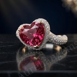 Könnte beinhalten: Ein Ring mit einem großen, herzförmigen, tiefroten Edelstein in einer goldfarbenen Metallschiene. Der Mittelstein ist von einem Kranz aus kleinen, klaren, funkelnden Steinen umgeben, wobei weitere kleine Steine auf der Ringschiene gefasst sind.