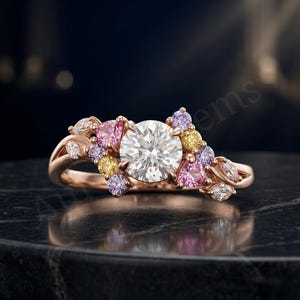 Könnte beinhalten: Ein Ring aus Roségold mit einem großen, runden, weißen Mittelstein, der von einer Ansammlung kleinerer rosa, gelber und lavendelfarbener Steine umgeben ist. Die Ringschiene ist seitlich mit weißen Steinen im Marquise-Schliff besetzt.