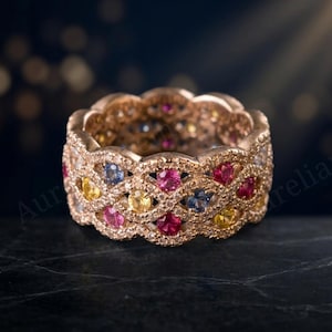 Könnte beinhalten: Ein Ring aus Roségold mit einem komplexen, geflochtenen Gitterdesign, das mit kleinen weißen Pavé-Steinen besetzt ist. Das Muster wird durch runde, mehrfarbige Edelsteine wie rosa, gelbe und blaue Saphire akzentuiert, die in das Metall gefasst sind.