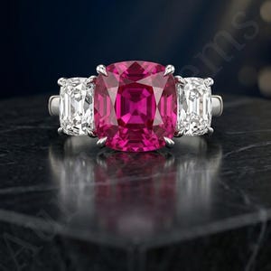 Puede incluir: Un anillo de tono plateado con una gran piedra preciosa de color rosa vibrante en talla cojín en el centro, flanqueada por dos piedras blancas transparentes rectangulares más pequeñas a cada lado. El engaste es de metal pulido con cuatro garras.