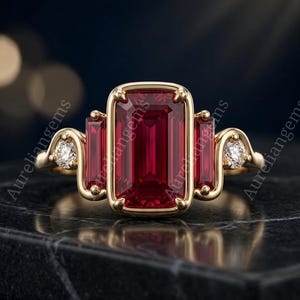 Peut inclure: Une bague en or ornée d'une grande pierre rouge taille émeraude au centre, flanquée de deux pierres rouges plus petites taille baguette et de deux diamants blancs ronds. L'anneau en métal a une finition lisse et le texte Aureliangems est visible.