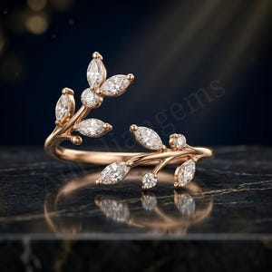 Pode incluir: Um anel aberto em ouro rosa com design de ramo e pedras brancas em lapidação marquise e redonda. A banda divide-se em dois caules delicados, cada um adornado com pedras em forma de folha e pequenas pedras redondas, criando um motivo botânico.