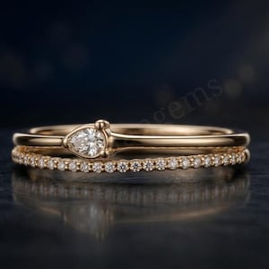 May include: A double-band ring crafted from polished gold. The top band features a single pear-shaped diamond set horizontally, while the bottom band is a delicate eternity style ring encrusted with a row of small, round, clear white diamonds.