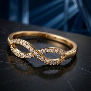 Puede incluir: Un anillo de oro con un diseño de símbolo de infinito en la parte frontal. El bucle del infinito está incrustado con pequeñas piedras claras de corte redondo que captan la luz. La banda es lisa y pulida, creando un aspecto continuo y refinado.
