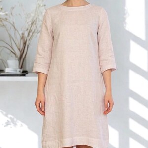 May include: A light pink linen dress with three-quarter sleeves. The dress has a simple, round neckline and falls to the knees. The text "FOF FUTURE OF FASHION" is in the upper left corner. The model has short, wavy hair.