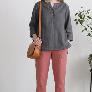 Può includere: Una donna indossa un top di lino grigio con maniche a tre quarti e scollo a V, abbinato a pantaloni di lino color corallo. Porta una borsa a tracolla in pelle marrone e tacchi abbinati. L'outfit è semplice ed elegante.
