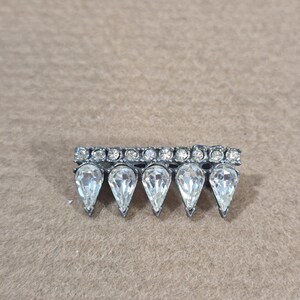 May include: A silver-toned metal brooch featuring a horizontal row of small, round, clear rhinestones above five larger, teardrop-shaped, clear rhinestones arranged in a downward-pointing, spiked pattern across the base of the piece.