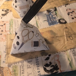 May include: Two white triangular pin cushions with black elastic bands. The pin cushions are decorated with sewing-themed patterns, including scissors, spools of thread, and buttons. The background features a fabric with vintage farm-themed designs and text.