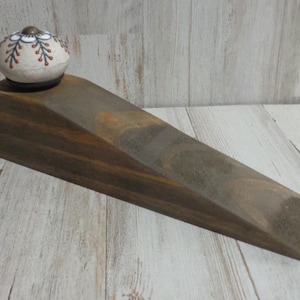 May include: A wooden doorstop with a decorative, round, ceramic top. The doorstop is a dark brown color and has a wedge shape. The ceramic top is white with a brown and blue floral design.
