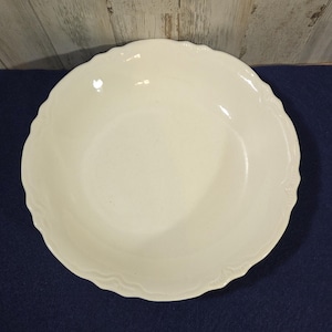 May include: A round, off-white ceramic bowl featuring a smooth, glossy finish and a decorative scalloped edge. The bowl has a wide, shallow basin and a simple, clean design, showcasing a uniform cream color throughout the entire piece.